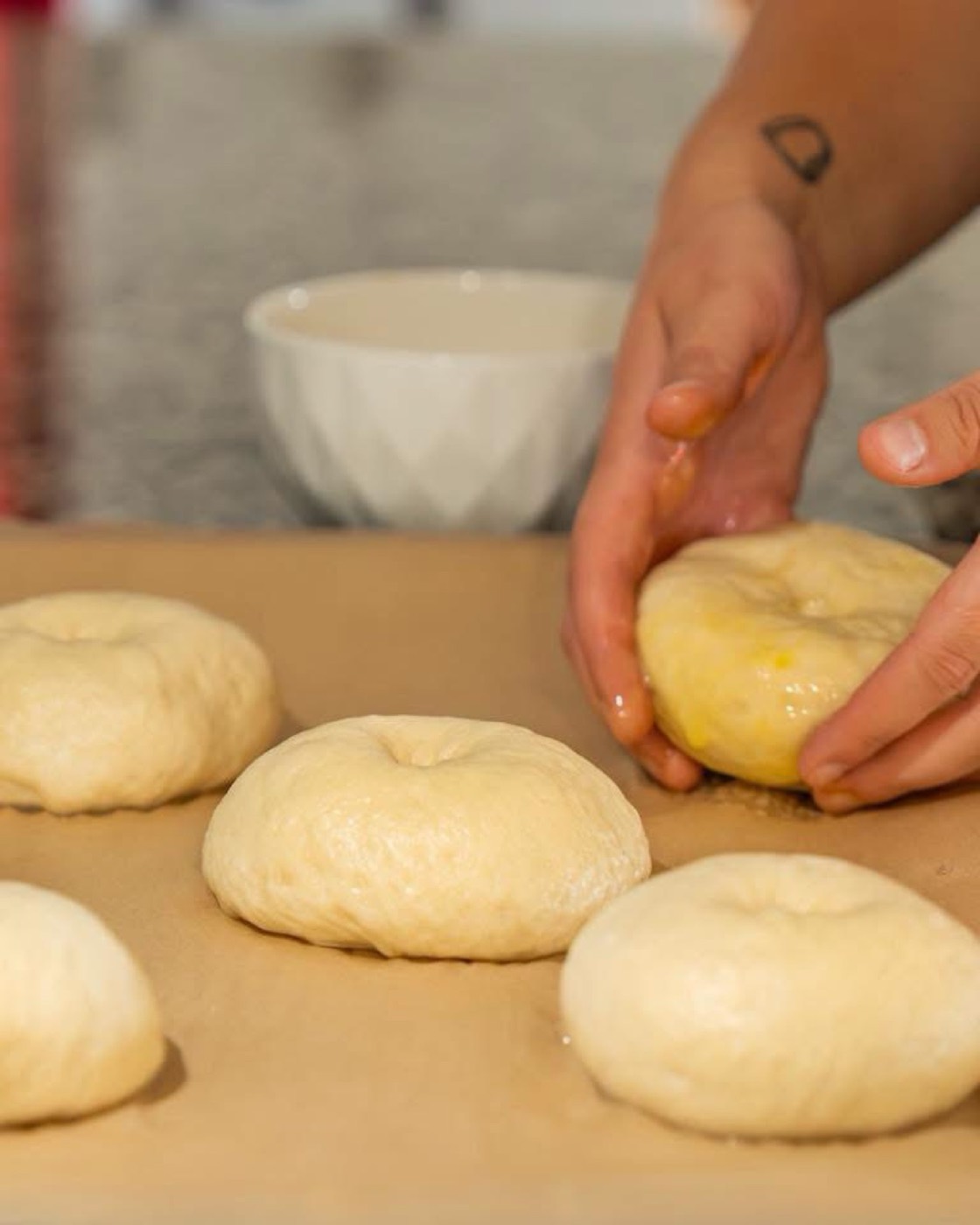 Hands brushing egg wash onto rounds of bagel dough on parchment