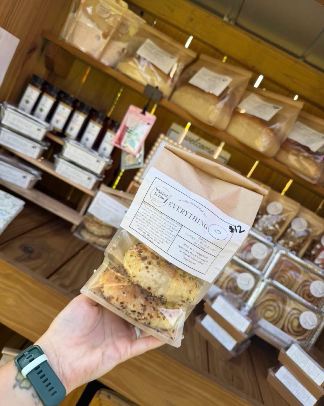 A kraft bag of Everything sourdough bagels held up in front of the stand shelves